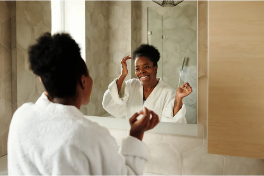 Mulher vestindo roupão branco admira no espelho do banheiro os efeitos da radiofrequência na pele do seu rosto.