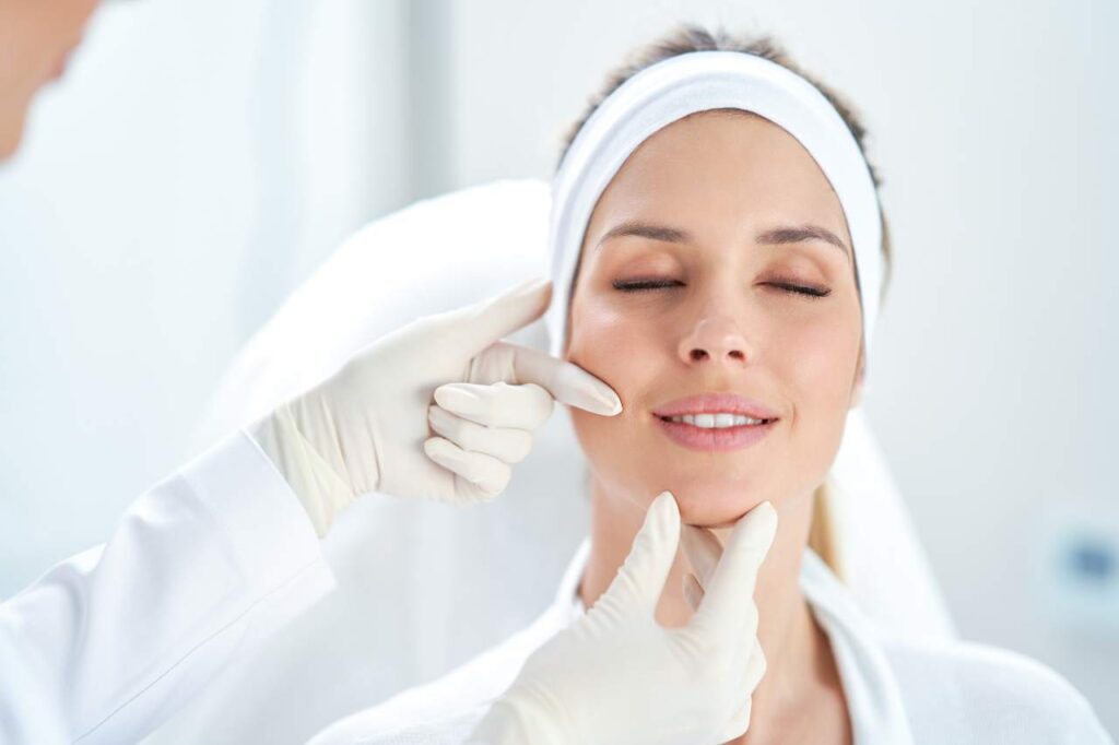 Mulher de olhos fechados sorrindo e profissional com luvas avaliando sua pele.