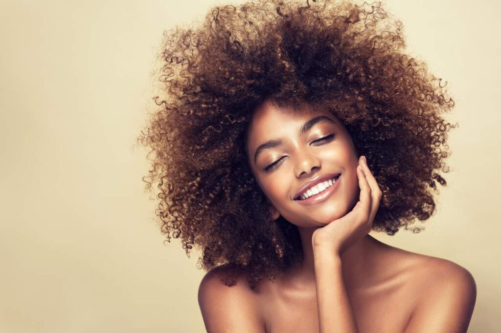 Mulher sorrindo de olhos fechados, com a cabeça levemente inclinada para o lado, apoiando o queixo em uma das mãos