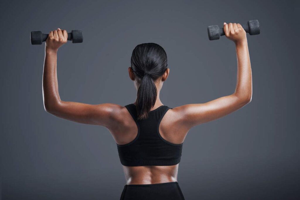 Mulher de costas, com roupas de academia, segurando halteres enquanto realiza exercício para os ombros.