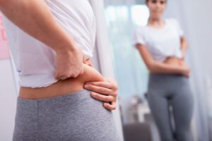 Mulher em frente ao espelho, pinçando sua barriga na lateral com as mãos, avaliando se há flacidez.