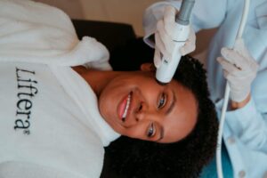 Mulher deitada sorrindo e profissional realizando um tratamento anti-idade com o ultrassom microfocado Liftera em sua face.